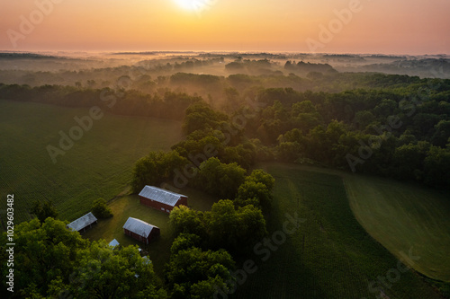 Foggy Iowa Sunrise With A Red Painted Barn and Homestead With Green Fields And Forest Stretching Into The Distance