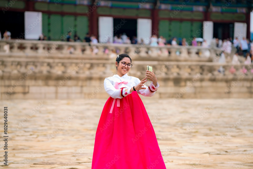 Naklejka premium Happy young Latina wearing a pink and white hanbok, standing in front of Gyeongbokgung Palace, taking selfie in Seoul, South Korea.