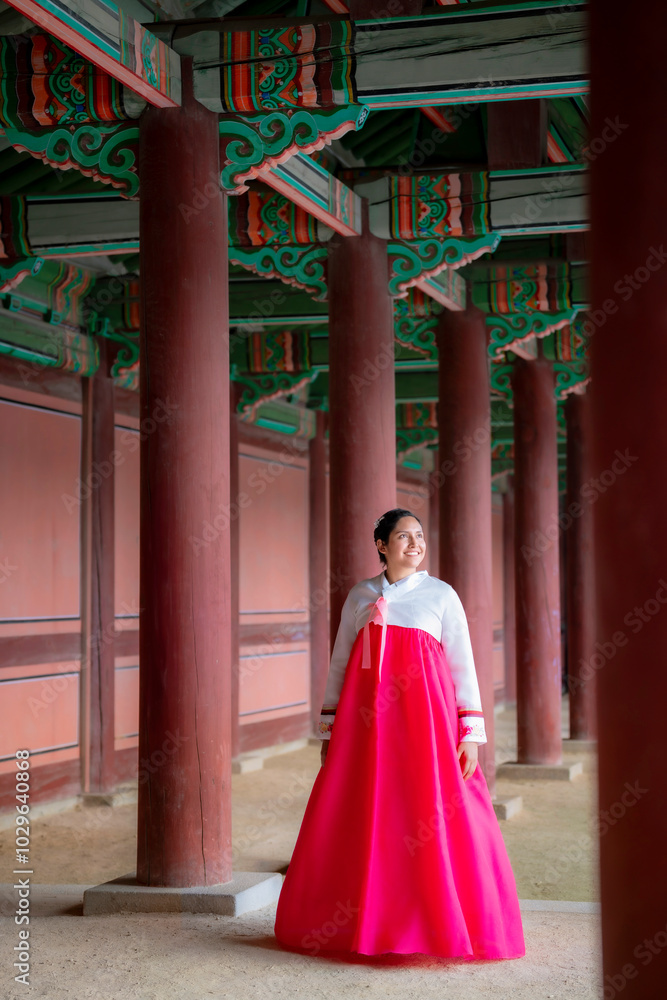 Young woman wearing a traditional pink hanbok stands under ornate wooden columns at Gyeongbokgung Palace in Seoul, South Korea. Young woman wearing a traditional pink hanbok stands under ornate wooden columns at Gyeongbokgung Palace in Seoul, South Korea.