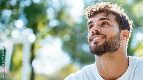 A close-up of a young man with curly hair and a beard, looking up with a smile against a blurred natural background, suggesting contemplation and optimism.