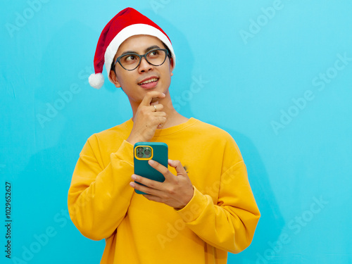 Asian man wearing christmas hat holding mobile phone showing thinking gesture with his eyes looking side up. photo