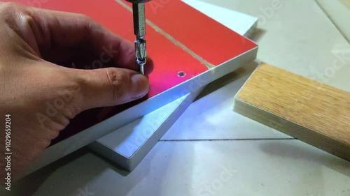 a man's hand installing a minifix screw into a red wooden board using a electric screwdriver drill machine. photo