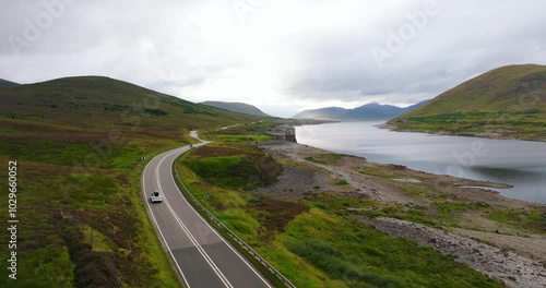 Aerial view of white electro car driving through epic landscape of Scottish hills and fields near cinematic calm lake