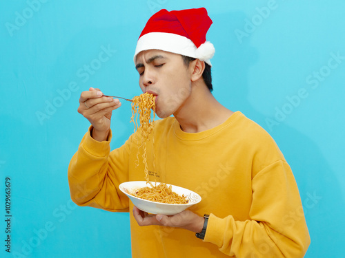 A happy Asian man wearing a yellow sweater and Christmas hat enjoying fried noodles with eyes closed, isolated on blue background. Perfect for festive, culinary, or lifestyle themes. photo