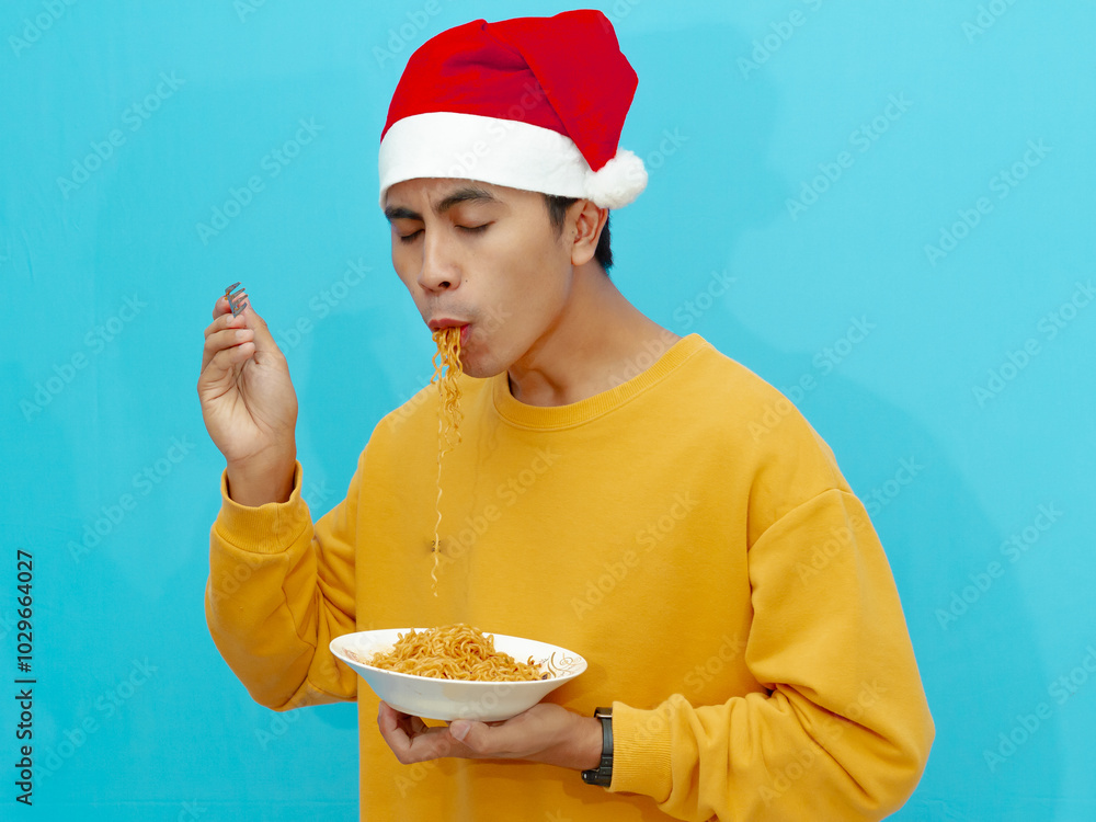 A cheerful Asian man wearing a yellow sweater and a Christmas hat enjoys eating fried instant noodles, isolated against a blue background. A cheerful Asian man wearing a yellow sweater and a Christmas hat enjoys eating fried instant noodles, isolated against a blue background.