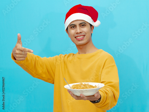 A cheerful Asian man wearing a yellow sweater and a Christmas hat, holding a plate of fried noodles and smiling while pointing, isolated against a blue background. photo