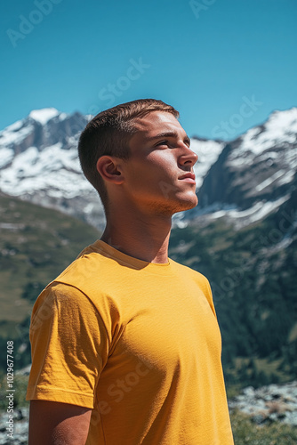 A man with short hair in a blank yellow t-shirt, standing in front of a mountain range, clear blue sky, side view, adventurous spirit.