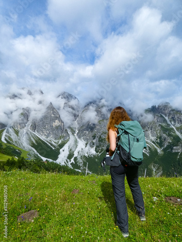 A female hiker stands with a backpack, facing and admiring a stunning mountainous landscape in the Tyrolean Alps