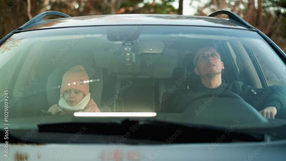 a man in a green jacket sits in the driver s seat of a car, while his daughter, wearing a pink beanie and jacket, is absorbed in her smartphone a man in a green jacket sits in the driver s seat of a car, while his daughter, wearing a pink beanie and jacket, is absorbed in her smartphone