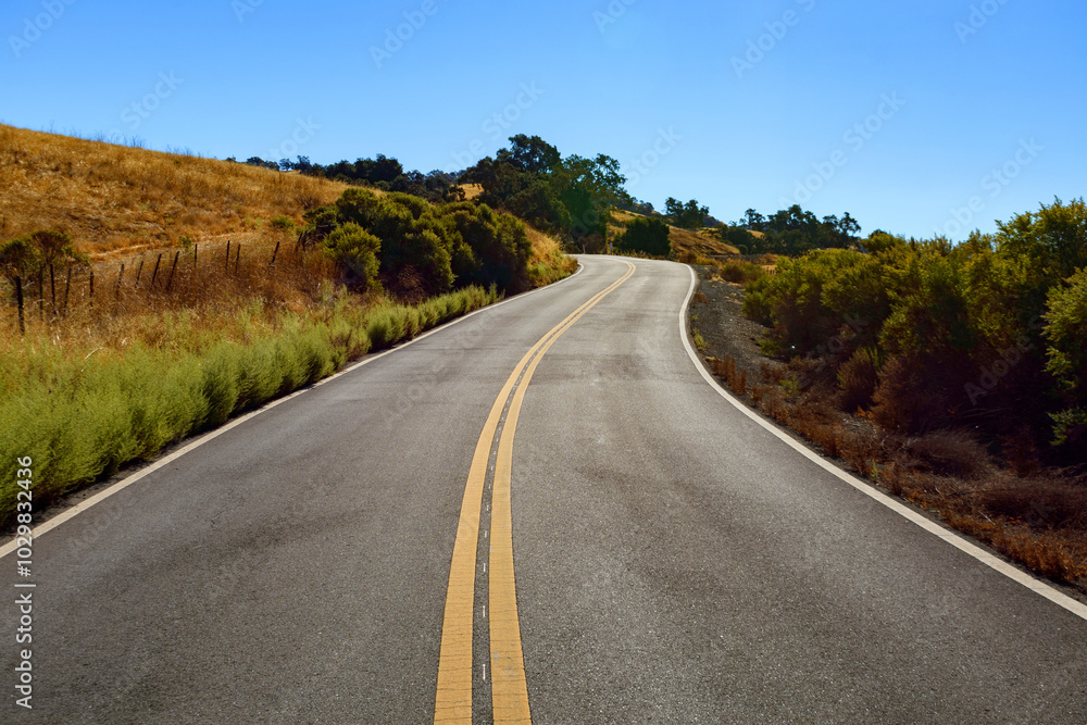 Mt. Hamilton Road winds through a dry, rural landscape with grassy hills and bushes under a clear blue sky Mt. Hamilton Road winds through a dry, rural landscape with grassy hills and bushes under a clear blue sky