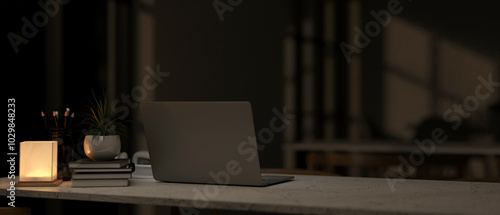 A back view of a laptop on a table in a modern dark room at nighttime, illuminated by a table lamp.