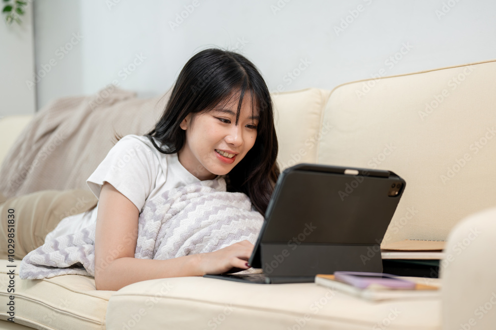 A happy Asian woman is using her digital tablet while laying on a sofa in the living room. A happy Asian woman is using her digital tablet while laying on a sofa in the living room.
