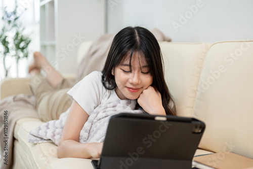 A happy Asian woman is using her digital tablet while laying on a sofa in the living room.