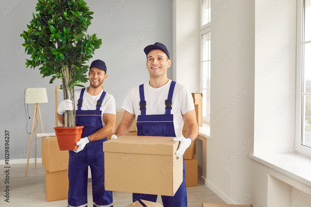 Two young smiling movers with plant and box in moving company client's new house. Caucasian male workers of moving service, dressed in work overalls, carry client's belongings into new apartment. Two young smiling movers with plant and box in moving company client's new house. Caucasian male workers of moving service, dressed in work overalls, carry client's belongings into new apartment.