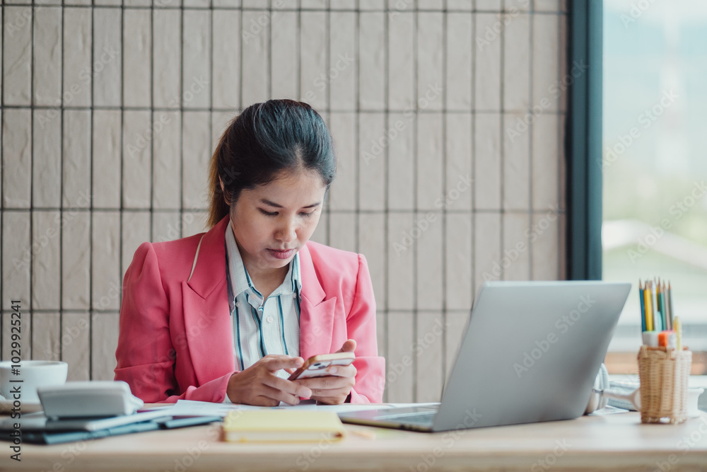 Professional woman in a pink blazer focused on her smartphone while working at a modern office desk with a laptop and stationery. Professional woman in a pink blazer focused on her smartphone while working at a modern office desk with a laptop and stationery.