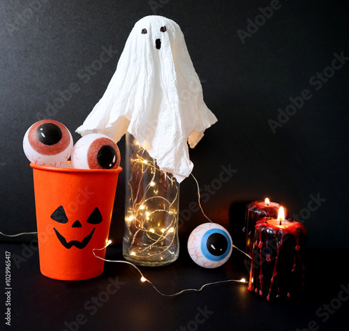 Photo of handmade decorations for Halloween. A Jack o lantern glass is filled with eyes. A ghost on a glass with a garland. Painted round eyes. Black candles with red drops. Black background. photo