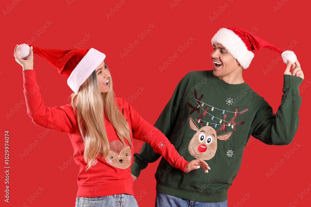 Young couple in Santa hats and Christmas sweaters on red background Young couple in Santa hats and Christmas sweaters on red background