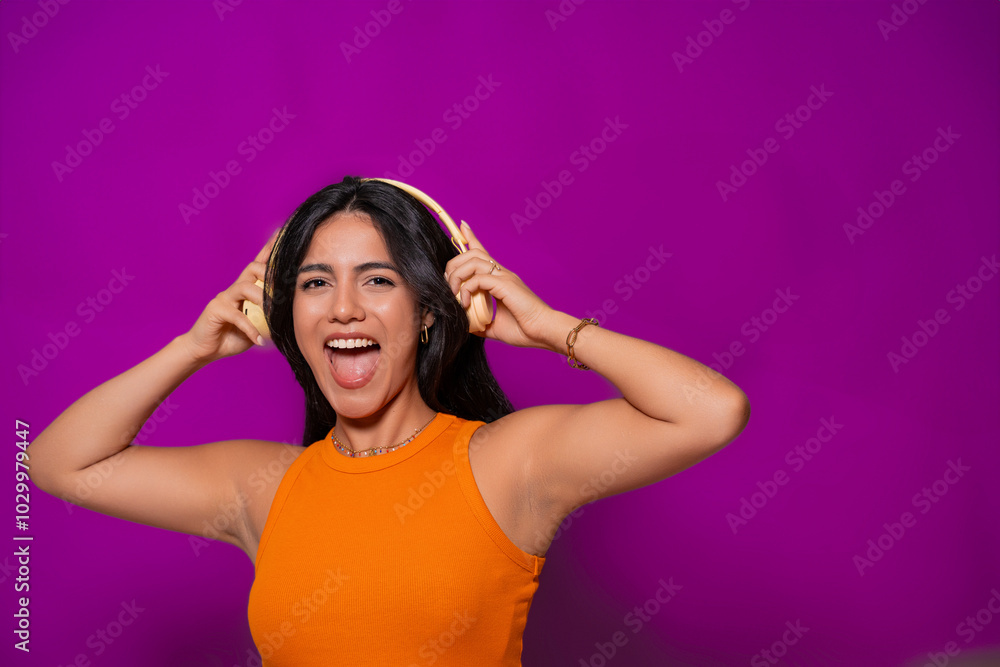Joyful woman in an orange top enjoying music with yellow headphones, smiling and making a fun expression against a vibrant purple background. Joyful woman in an orange top enjoying music with yellow headphones, smiling and making a fun expression against a vibrant purple background.