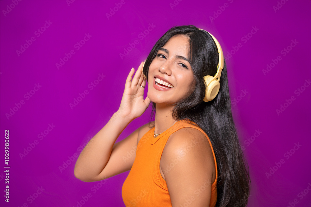 Side view of a smiling woman with yellow headphones, wearing an orange top, enjoying music and posing playfully against a purple background. Side view of a smiling woman with yellow headphones, wearing an orange top, enjoying music and posing playfully against a purple background.