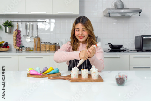 Young asian woman in an apron using a piping bag, pipe the icing onto the cupcake, decorating cupcake on a wooden board, adding a colorful touch to the cupcakes' presentation. photo