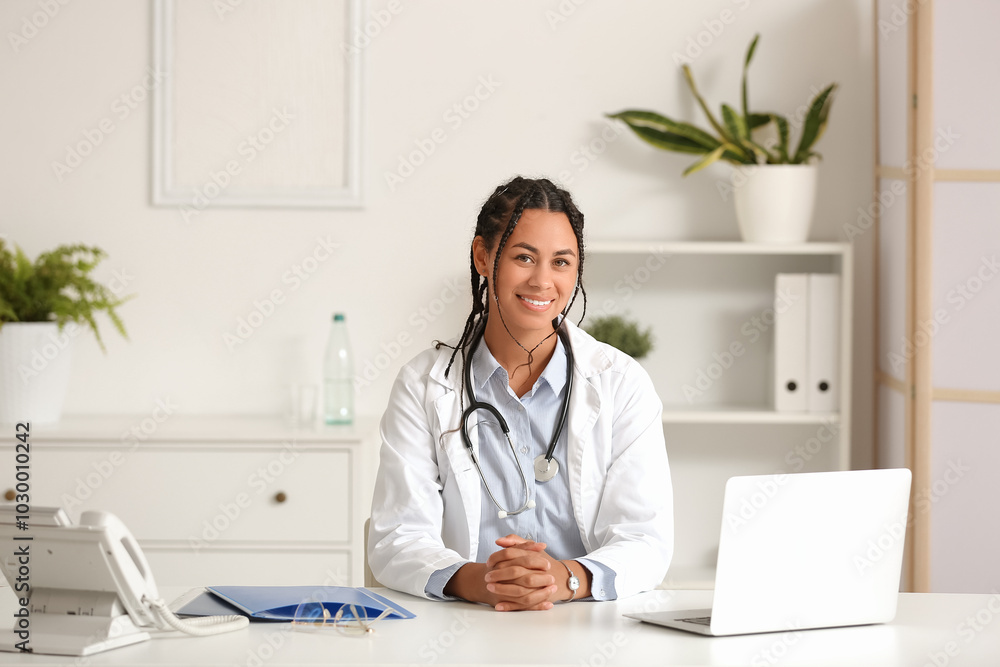Young African-American female doctor with telephone and laptop sitting at table in clinic Young African-American female doctor with telephone and laptop sitting at table in clinic