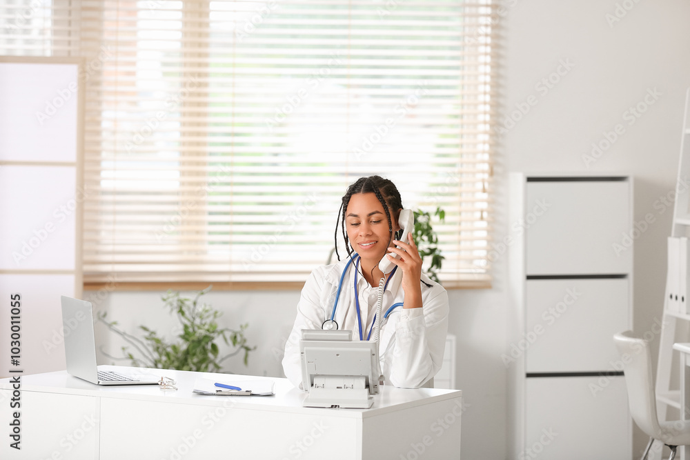 Young African-American female doctor with telephone working at table in clinic Young African-American female doctor with telephone working at table in clinic