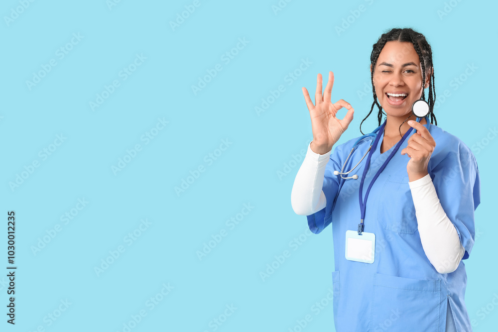 Young African-American female doctor with stethoscope and badge showing OK gesture on blue background Young African-American female doctor with stethoscope and badge showing OK gesture on blue background