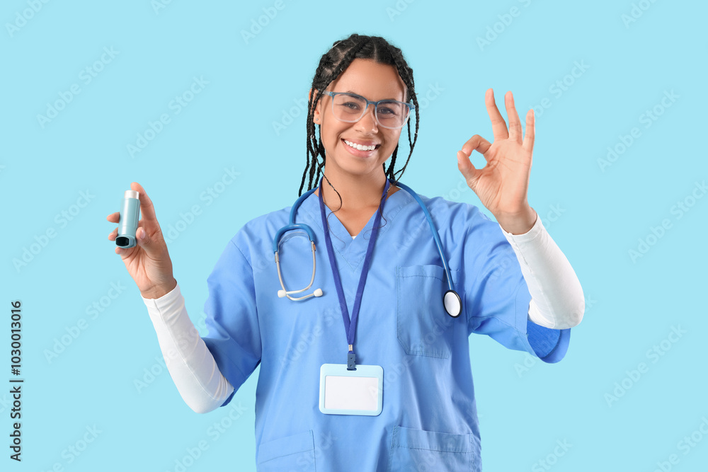 Young African-American female doctor with stethoscope and asthma inhaler showing OK gesture on blue background Young African-American female doctor with stethoscope and asthma inhaler showing OK gesture on blue background