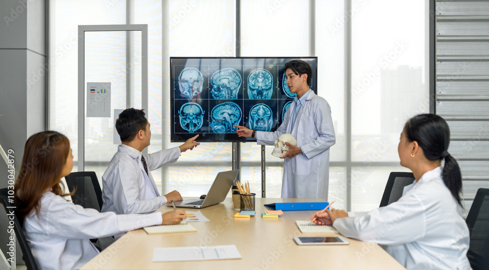 Obraz premium Young medical professional standing and presenting, holding a model of human skull and referencing image of brain scan displayed on a screen. The audience listening attentively with the presentation.