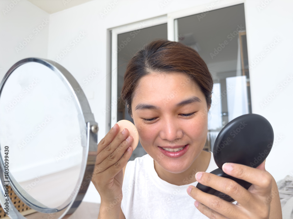 Young adult asian Thai woman wearing make up, using puff applying powder on her face and look at mirror, morning routine beauty concept. Young adult asian Thai woman wearing make up, using puff applying powder on her face and look at mirror, morning routine beauty concept.