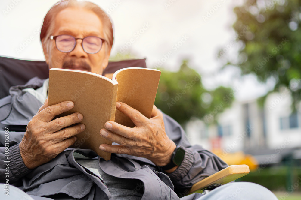 Elderly Asian man with eyeglasses reading a book outdoors in autumn by a tree, looking thoughtful. Elderly Asian man with eyeglasses reading a book outdoors in autumn by a tree, looking thoughtful.