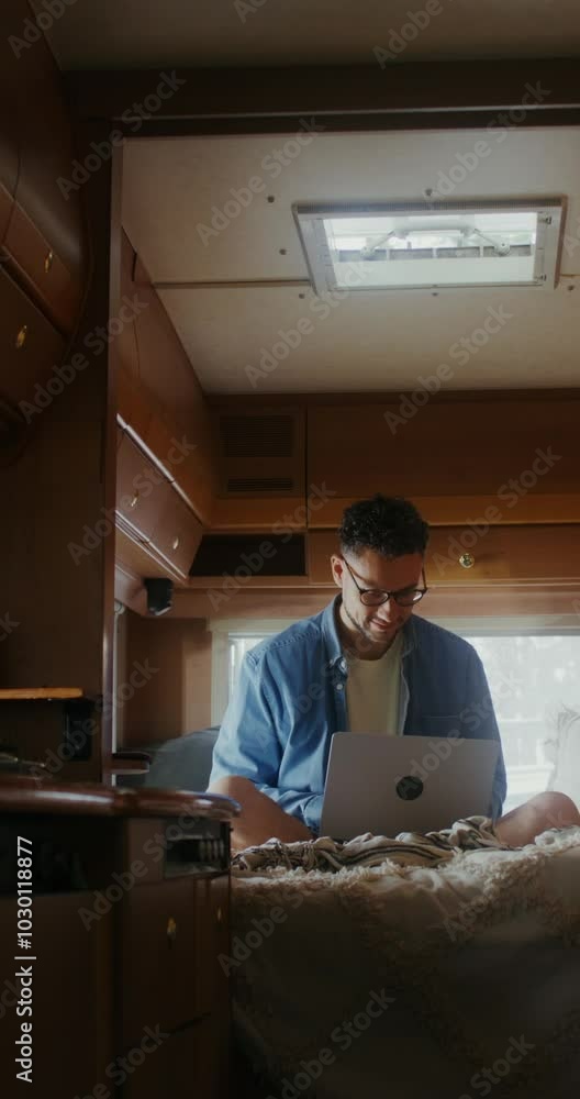 A young man with glasses uses a laptop sitting on a bed in a van. The woman smiles as she sits down next to him A young man with glasses uses a laptop sitting on a bed in a van. The woman smiles as she sits down next to him