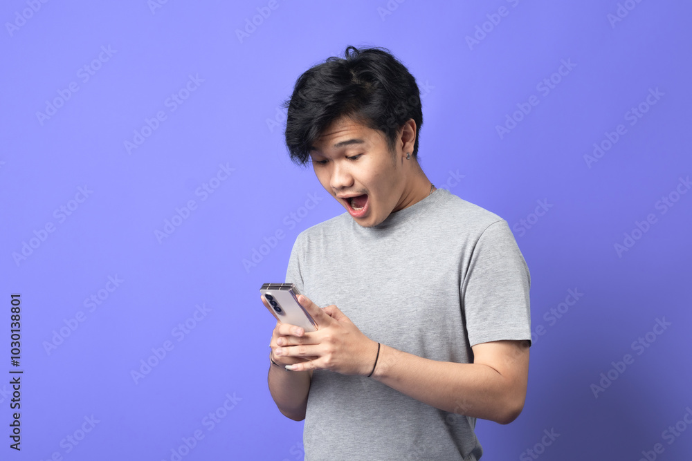 A young Asian man wearing a gray t-shirt is looking at his smartphone with an expression of surprise or excitement. He stands against a solid purple background, holding the phone with both hands A young Asian man wearing a gray t-shirt is looking at his smartphone with an expression of surprise or excitement. He stands against a solid purple background, holding the phone with both hands