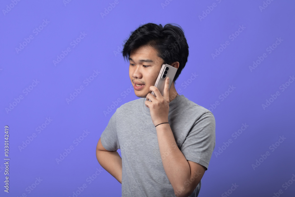 A young Asian man in a gray t-shirt is standing against a purple background, holding a smartphone to his ear as if engaged in a conversation. He has a casual stance with one hand on his hip A young Asian man in a gray t-shirt is standing against a purple background, holding a smartphone to his ear as if engaged in a conversation. He has a casual stance with one hand on his hip