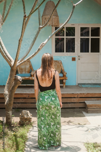 Back view of a young blonde woman in swimsuit and cape with tropical print standing in front of a thatched roof bungalow during her vacation. Island hotel on ocean shore.