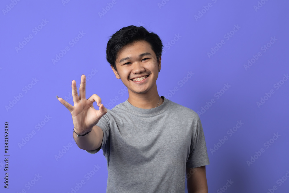 A young Asian man is smiling, standing in a studio with a purple background. He is wearing a gray t-shirt and making an A young Asian man is smiling, standing in a studio with a purple background. He is wearing a gray t-shirt and making an
