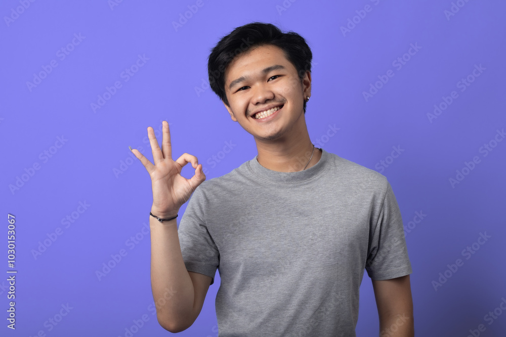 A young Asian man is smiling, standing in a studio with a purple background. He is wearing a gray t-shirt and making an A young Asian man is smiling, standing in a studio with a purple background. He is wearing a gray t-shirt and making an