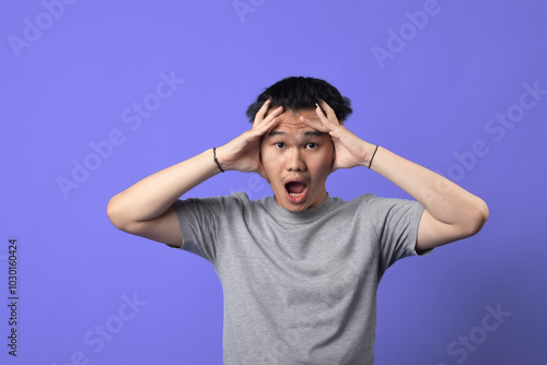 Asian man is seen in a moment of frustration, pulling his hair with both hands while clenching his teeth and shutting his eyes tightly. wearing a gray t-shirt and is set against a purple background