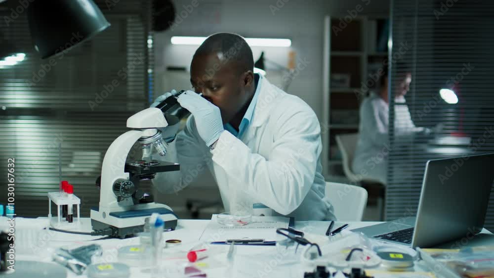 Black forensic scientist examining evidence under microscope and taking notes at desk in crime lab Black forensic scientist examining evidence under microscope and taking notes at desk in crime lab