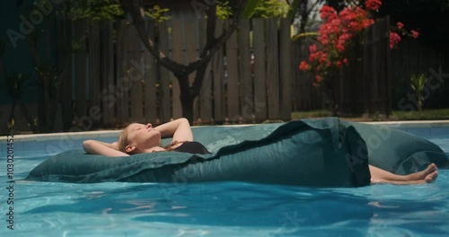 Young woman floating on a mattress in a pool with her eyes closed and enjoying her vacation on a tropical island. Relaxation and wellness treatments at all inclusive hotel. Spa treatments and swimming