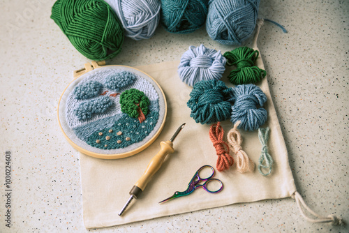 Beautiful embroidered landscape in a frame placed on piece of fabric together with woolen skeins, punch needle and scissors