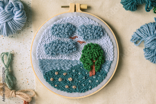 Top view of beautiful embroidered landscape in green and blue colors in beige frame surrounded with woolen yarns of matching colors.