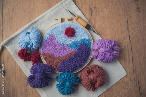 Top view of embroidered mountain landscape in violet colors surrounded by skeins of yarn and a punch needle.
