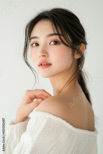 Confident Asian woman touching her shoulder, posed in bright pro studio, showcasing minimalistic fashion look against simple white background