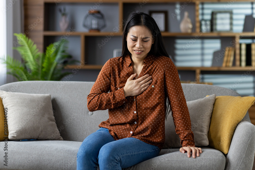 Asian woman expressing pain while seated on sofa, clutching chest in modern living room. Emotions of discomfort and concern are evident in her expression. Asian woman expressing pain while seated on sofa, clutching chest in modern living room. Emotions of discomfort and concern are evident in her expression.