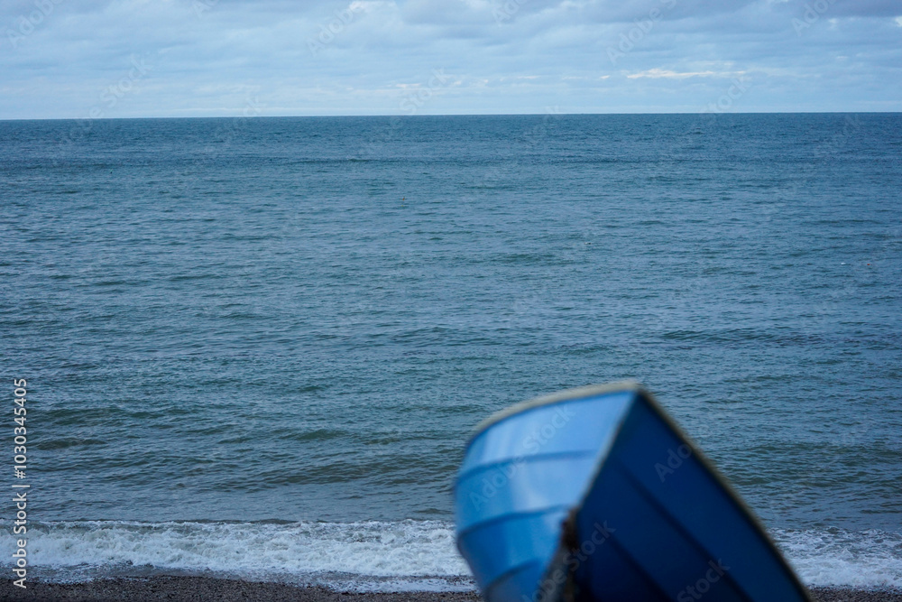 A blue boat sits on the shore, partially visible, with the vast ocean extending toward the horizon under a cloudy sky A blue boat sits on the shore, partially visible, with the vast ocean extending toward the horizon under a cloudy sky