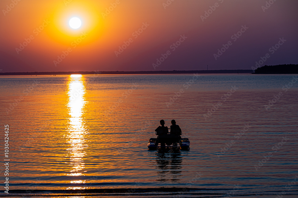 couple of young people are sitting on catamaran on background of multicolored sunset sky, sun disk and sunset track on the water couple of young people are sitting on catamaran on background of multicolored sunset sky, sun disk and sunset track on the water