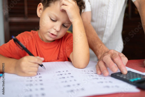 A boy does his math homework while his father stands next to him, helping him check the calculations using a calculator.