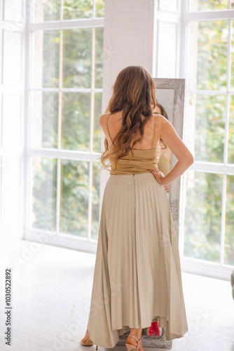 Beautiful Caucasian girl looks in . mirror in a beige top and a milk-colored maxi skirt, view from the back. Long curls. Women. Fashion and beauty concept Free Download Beautiful Caucasian girl looks in . mirror in a beige top and a milk-colored maxi skirt, view from the back. Long curls. Women. Fashion and beauty concept - GFXLoad