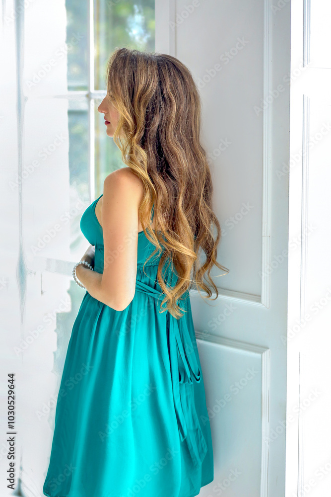 Attractive beautiful brunette girl with curls and blue blowouts in a green midi dress. Light interior room with green flowers. Attractive beautiful brunette girl with curls and blue blowouts in a green midi dress. Light interior room with green flowers.
